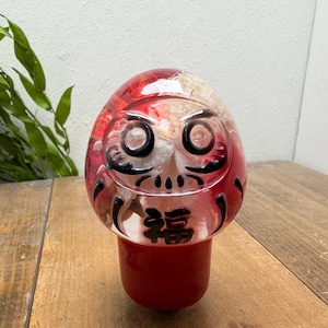 Op de afbeelding: Een helder, afgerond Daruma-popfiguur met een rode basis en een transparant bovendeel gevuld met gedroogde bloemen. De pop heeft zwart geschilderde gelaatstrekken, waaronder dikke wenkbrauwen en een snor, met het zwarte Kanji-teken 福 op de voorzijde.