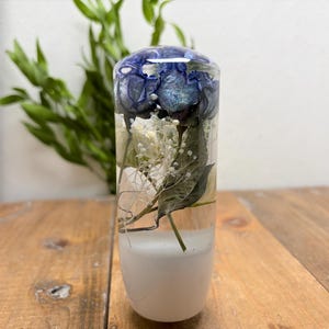 May include: A clear, cylindrical resin object with a white base, encasing blue and white flowers and greenery. The top of the object has a dark blue, swirled design. The object is displayed on a wooden surface.