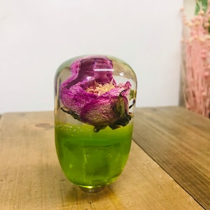 Peut inclure: Un dôme en verre transparent contenant une fleur violette séchée. Le dôme est rempli d'un liquide vert.