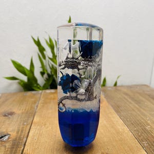 Könnte beinhalten: Klarer, zylindrischer Gegenstand aus Kunstharz mit einer detaillierten silbernen Drachenskulptur. Die untere Hälfte ist tiefblau, geht in transparent mit blauen Akzenten über. Der Drache ist von blauen und weißen Details umgeben.