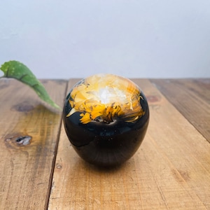 Op de afbeelding: Een zwarte harsbol met een gele en witte bloem erin ingekapseld. De bol ligt op een houten oppervlak.