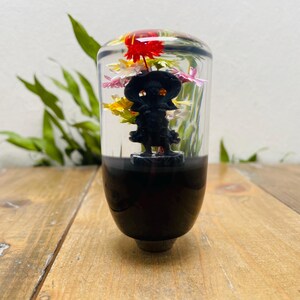 Puede incluir: Un pomo de palanca de cambios cilíndrico transparente con una base negra y una figura negra en el interior. El pomo está lleno de flores coloridas, incluyendo rojo, amarillo y rosa. El pomo está sobre una superficie de madera.