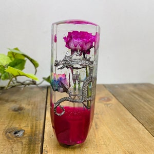 May include: A clear glass gear shift knob featuring a silver dragon design and a pink flower inside. The base of the knob is filled with pink resin.