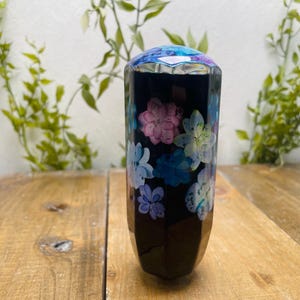 Puede incluir: Un jarrón negro facetado con un diseño floral en tonos rosa, azul y blanco. La parte superior del jarrón tiene un diseño geométrico multicolor. El jarrón se exhibe sobre una superficie de madera.