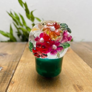 Puede incluir: Un tapón de botella decorativo de vidrio con un diseño floral. El tapón presenta una parte superior transparente y redondeada con un grupo de flores rojas, rosas y naranjas. La base es de color verde vibrante. El tapón está sobre una superficie de madera.