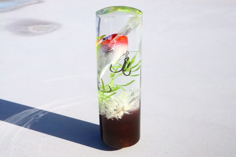 May include: A clear glass cylinder containing a white and red fishing lure, green plant, and a white flower. The base of the cylinder is filled with a dark brown liquid.