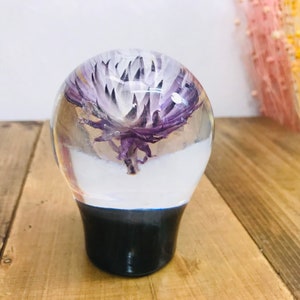 May include: A clear glass sphere with a purple and white flower inside. The sphere is sitting on a black base.