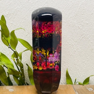 Puede incluir: Un tapón de botella decorativo con fondo negro y diseño floral en tonos rojos, rosas, amarillos y morados. El tapón tiene un acabado brillante y una base geométrica.