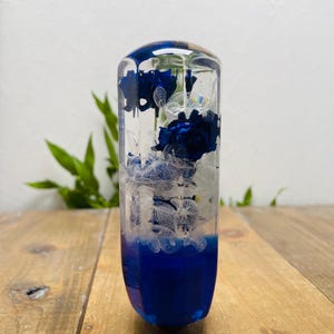 Könnte beinhalten: Ein klares, zylindrisches Kunstwerk aus Harz mit einem tiefblauen Sockel und eingebetteten blauen Rosen und weißen Blüten. Die Blumen sind in transparentem Harz eingeschlossen.