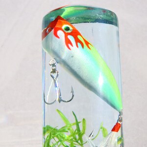 Pu&ograve; includere: Un'esca da pesca bianca e verde con fiamme rosse e arancioni &egrave; racchiusa in un cilindro trasparente pieno d'acqua e piante verdi.