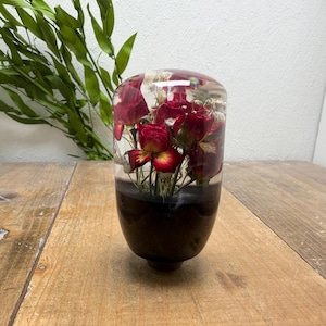 May include: A cylindrical gear shift knob featuring a clear resin top section embedded with dried red roses and small white flowers, transitioning into a solid black base. The object has a smooth, polished finish and a rounded top surface.