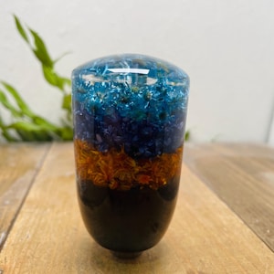 May include: A decorative resin object with a rounded top and a dark base. The top section is clear resin with blue and gold inclusions. The middle section is orange, and the base is black. It is on a wooden surface.