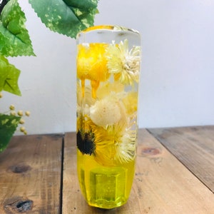 Op de afbeelding: Een transparante glazen cilinder gevuld met gedroogde gele en witte bloemen. De bloemen zijn in een willekeurig patroon gerangschikt en zijn zichtbaar door het heldere glas. De cilinder staat op een houten oppervlak.