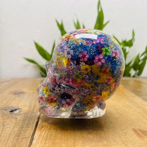 Könnte beinhalten: Ein klarer Kunstharzschädel gefüllt mit bunten Trockenblumen. Die Blumen sind eine Mischung aus Rosa, Blau, Gelb und Grün und bilden ein lebendiges Dekorationsstück. Der Schädel steht auf einer Holzoberfläche.