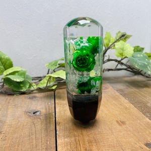 Könnte beinhalten: Ein grüner Rosenkopf, der in klarem Harz eingebettet ist, auf einem schwarzen Sockel montiert. Der Harz-Schalthebelknopf ist ein einzigartiges und dekoratives Autozubehör.