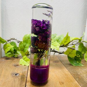 Può includere: Un cilindro di vetro trasparente riempito con fiori secchi viola e marroni, con una base viola. Il cilindro è appoggiato su una superficie di legno.