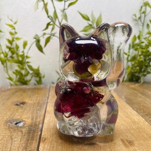 Könnte beinhalten: Klare Katzenfigur aus Kunstharz mit einem dunkelroten Blumenarrangement im Inneren. Die Katze sitzt mit erhobener Pfote. Sie steht auf einer Holzoberfläche, im Hintergrund ist grünes Laub zu sehen.