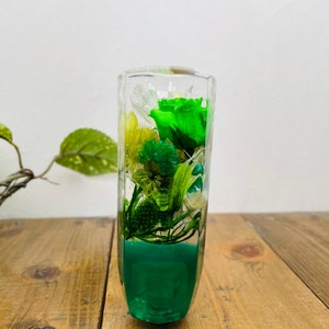 Op de afbeelding: Een transparante glazen cilinder gevuld met geconserveerde groene en witte bloemen. De bloemen zijn in een boeket gerangschikt en de cilinder is gevuld met een groene vloeistof.