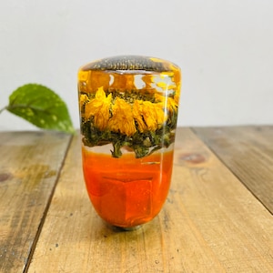 Op de afbeelding: Een transparante glazen pookknop met een laag gedroogde gele bloemen en groene bladeren ingekapseld in hars. De onderste laag is effen oranje.