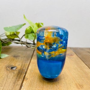 Op de afbeelding: Een blauwe en gele hars versnellingspookknop met geperste bloemen erin. De knop is rond en heeft een glad oppervlak.