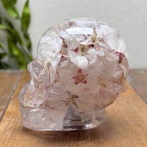 以下が含まれることがあります： 白い花と淡いピンクの花が埋め込まれた、頭蓋骨の形をした透明な樹脂製の彫刻。透明な素材の中に繊細な花びらと茎が浮かんでおり、詳細な解剖学的構造が表現されています。