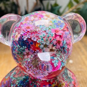 Könnte beinhalten: Eine transparente Harz-Bärenfigur, gefüllt mit gepressten Blumen in Rosa-, Blau- und Grüntönen. Der Bär hat eine glänzende Oberfläche und ist ein dekoratives Stück.