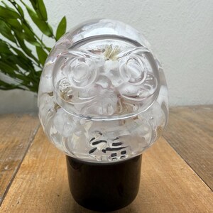 Könnte beinhalten: Ein klarer, kugelförmiger Briefbeschwerer aus Kunstharz mit einem eingebetteten Daruma-Puppendesign und weißen floralen Elementen im Inneren. Das Objekt hat eine glatte, abgerundete Oberseite und steht auf einem schwarzen zylindrischen Sockel.