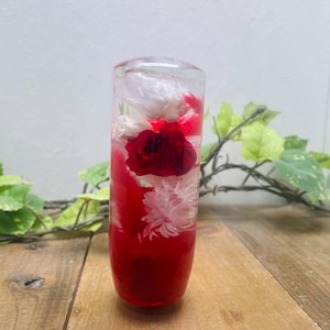 Può includere: Un oggetto decorativo cilindrico trasparente riempito con un liquido rosso vibrante e fiori assortiti. I fiori includono una rosa rossa prominente e fiori bianchi, creando un contrasto visivo sorprendente. L'oggetto è esposto su una superficie di legno.