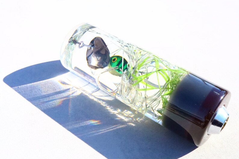 May include: A clear glass cylinder containing a green and black fishing lure. The cylinder is filled with clear liquid and has a dark brown base.