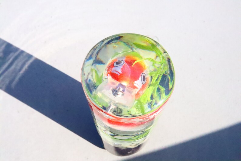 May include: A clear glass cylinder containing a red and white goldfish figurine. The cylinder is filled with water and green plastic plants.