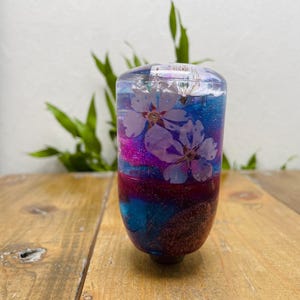 May include: A clear, cylindrical resin object with embedded white cherry blossoms. The object features layers of blue, pink, and maroon glitter, and sits on a wooden surface. The base is a dark brown colour.