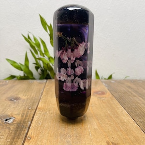 Puede incluir: Un pomo de palanca de cambios hexagonal de color púrpura oscuro con detalles florales rosas y blancos incrustados. El pomo está hecho de un material transparente, lo que permite que las flores sean visibles. Se encuentra sobre una superficie de madera.