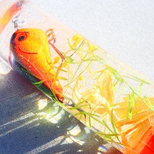 May include: A clear glass cylinder filled with a yellow and orange lure, green and yellow plant material, and a clear liquid. The cylinder has a red cap.