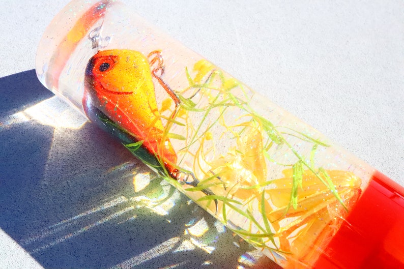 May include: A clear glass cylinder filled with a yellow and orange lure, green and yellow plant material, and a clear liquid. The cylinder has a red cap.