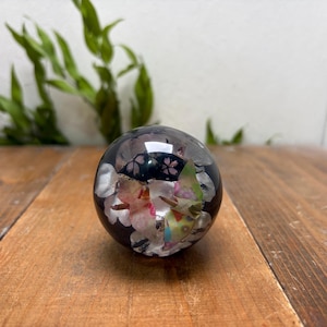 May include: A clear, spherical paperweight with a dark background, encasing colorful floral elements. The paperweight sits on a wooden surface, with green foliage in the background. The sphere reflects light, highlighting the embedded flowers.