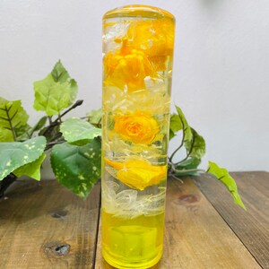 May include: A clear glass cylinder filled with yellow flowers and white petals. The cylinder has a yellow base.