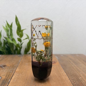 May include: A cylindrical gear shift knob made of clear resin, featuring embedded dried yellow flowers and green stems. The base of the knob is a solid dark purple color, and the top is rounded. The object has a smooth, polished finish throughout.