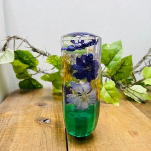 Op de afbeelding: Een transparante glazen pookknop met een groene basis en geperste bloemen erin. De bloemen zijn paars, blauw, geel en wit.