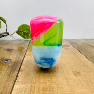 Può includere: Un pomello del cambio cilindrico e colorato con un design marmorizzato. La sezione superiore è rosa brillante, che sfuma in verde, poi in blu e bianco alla base. Il pomello è appoggiato su una superficie di legno.