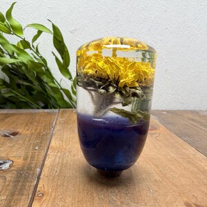 Könnte beinhalten: Ein zylindrischer Schaltknauf aus klarem Kunstharz mit einer konservierten gelben Blume im oberen Bereich und einer tiefvioletten, undurchsichtigen Basis. Das Objekt hat eine glatte, polierte Oberfläche und eine abgerundete Oberseite.