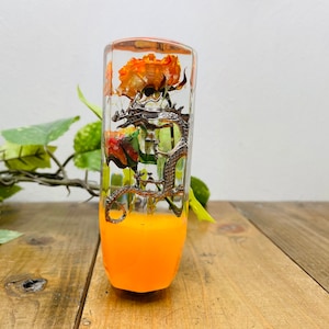 Op de afbeelding: Een transparante resin versnellingspookknop met een zilveren draakontwerp en oranje bloemen erin. De knop heeft een fel oranje basis.