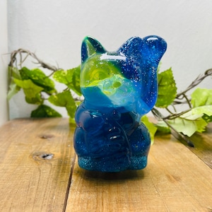 May include: A resin cat figurine in shades of blue, green, and yellow. The cat has a glossy finish with glitter accents. The figurine is sitting on a wooden surface, with greenery in the background.