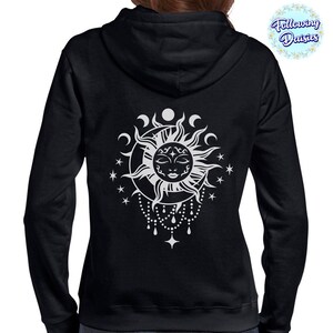 K&ouml;nnte beinhalten: Schwarzer Kapuzenpullover mit einem wei&szlig;en Sonnen- und Mondmotiv. Die Sonne hat ein Gesicht und ist von Sternen und Mondsicheln umgeben. Das Design h&auml;ngt an einer Perlenkette.