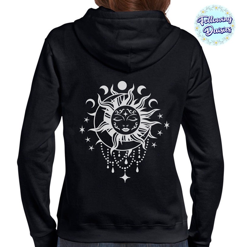 K&ouml;nnte beinhalten: Schwarzer Kapuzenpullover mit einem wei&szlig;en Sonnen- und Mondmotiv. Die Sonne hat ein Gesicht und ist von Sternen und Mondsicheln umgeben. Das Design h&auml;ngt an einer Perlenkette.