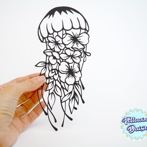 May include: Black paper cut-out of a jellyfish with flowers and leaves. The jellyfish is in the center of the image and is surrounded by flowers and leaves. The image is on a white background.