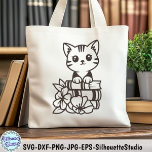 Puede incluir: Una bolsa de tela blanca con una ilustración en blanco y negro de un gato sentado sobre una pila de libros. El gato mira al espectador y tiene una expresión linda. Los libros están apilados uno encima del otro y tienen un diseño simple. También hay flores blancas y negras en la bolsa.