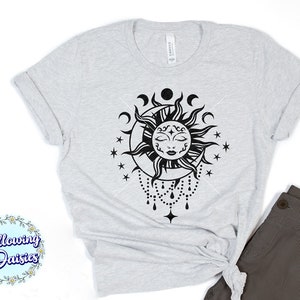 K&ouml;nnte beinhalten: Ein graues T-Shirt mit einem schwarzen und wei&szlig;en Sonnen- und Monddesign. Die Sonne hat ein Gesicht mit geschlossenen Augen und ein Halbmond befindet sich dahinter. Es gibt Sterne und eine Perlenkette, die vom unteren Rand des Designs h&auml;ngt.