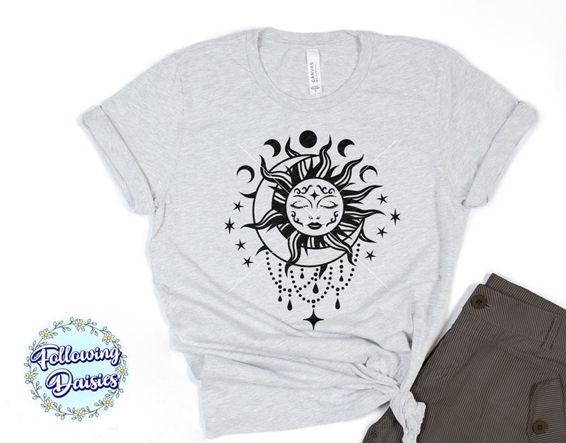 K&ouml;nnte beinhalten: Ein graues T-Shirt mit einem schwarzen und wei&szlig;en Sonnen- und Monddesign. Die Sonne hat ein Gesicht mit geschlossenen Augen und ein Halbmond befindet sich dahinter. Es gibt Sterne und eine Perlenkette, die vom unteren Rand des Designs h&auml;ngt.