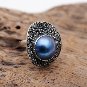 May include: A silver ring featuring a large, iridescent blue pearl. The pearl is set in a circular frame covered in small, dark gray beads. The ring is displayed on a textured, light brown wooden surface.
