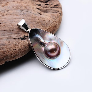 Peut inclure: Un pendentif en forme de goutte d'eau avec une bélière argentée. Le pendentif présente une grande coquille irisée avec une perle brun foncé au centre. La coquille a une surface chatoyante et multicolore. Le pendentif est posé sur un fond en bois.